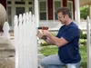 A man is kneeling and using a cordless drill to repair a white picket fence. A house is visible in the background. Podgląd zdjęcia produktu