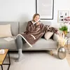 A woman is sitting on a couch wearing a brown hoodie blanket. Produktbild-Vorschau