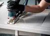 A person wearing black gloves is using a jigsaw to cut a white surface, with sawdust flying. Voir les images du produit