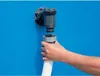 A hand is shown connecting a white hose to a gray filter pump attached to a blue surface, likely a swimming pool wall. Voir les images du produit