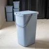 A light blue trash can with a lid and handle is shown in a kitchen or utility room setting, with other similar trash cans in the background. Produktbild-Vorschau