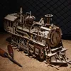 A wooden model of a steam locomotive sits on a surface covered in wood shavings, with two woodworking tools in the foreground. Náhľad obrázku produktu