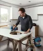 A man is using a Bosch jigsaw to cut a circular hole in a countertop, with a Bosch vacuum cleaner nearby. Produktbild-Vorschau