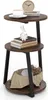 A three-tiered round side table with a rustic brown wood finish and black metal legs, decorated with a coffee cup, books, a ceramic rabbit figurine, and decorative items in a basket. Podgląd zdjęcia produktu