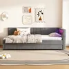 A gray upholstered daybed with pillows and a blanket, set against a white wall with framed art and a wall lamp. A rug with a floral accent is on the floor in front of the bed. Podgląd zdjęcia produktu