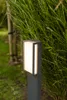 A modern outdoor LED pathway light with a dark gray metal finish and a frosted white diffuser, set against a backdrop of lush green grass and foliage. Anteprima dell’immagine prodotto