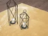 Two black metal lanterns with white candles inside, placed on a soft, beige rug next to a wooden floor. Produktbild-Vorschau