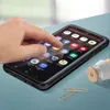 A hand is shown interacting with a smartphone that is placed on a blue surface. The phone is in a black case and displays various app icons on its screen. A paperclip and a small wooden object are also visible on the surface. Podgląd zdjęcia produktu