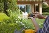 A person is holding a battery for a hedge trimmer in a garden setting. Produktbild-Vorschau