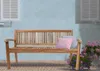 A wooden garden bench with two cushions sits on a brick patio in front of a light blue wall with white shutters. Produktbild-Vorschau