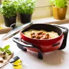 A red electric skillet filled with spaghetti and meatballs, placed on a kitchen counter with basil plants and garlic in the background. Produktbild-Vorschau