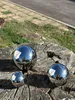 Three polished stainless steel garden spheres of different sizes are placed on a weathered tree stump outdoors, with a paved path and grass in the background. Produktbild-Vorschau