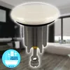 A close-up of a white ceramic drain stopper with a gray plastic base and a yellow adjustment nut, set against a blurred background of a modern bathroom. Produktbild-Vorschau