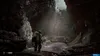 A lone figure in a futuristic suit walks through a dark, cavernous environment with strange glowing pools on the ground. Produktbild-Vorschau