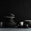A collection of dark-colored ceramic dishes and cups are stacked and arranged on a dark surface against a black background. Produktbild-Vorschau