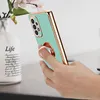 A hand holding a turquoise phone case with a gold trim and a rose gold ring holder, next to a vase with flowers. Náhľad obrázku produktu