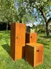 Three weathered metal pedestals of different sizes (labeled L, M, and S) are arranged in a grassy garden setting. A small planter with white flowers sits on top of the 'S' pedestal. Trees and greenery form the background. Produktbild-Vorschau