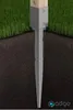 A metal post anchor is driven into the ground, securing a wooden post. The image shows the anchor partially buried in soil, with grass above. Anteprima dell’immagine prodotto