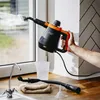 A hand holding a steam cleaner and spraying steam onto a window, with cleaning accessories on a countertop. Podgląd zdjęcia produktu