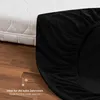 A close-up of a black fitted sheet on a mattress, with a portion of the mattress visible. The image is set against a wooden floor. Produktbild-Vorschau