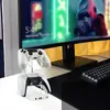 Two white game controllers are displayed on a clear stand on a desk in front of a computer monitor. Produktbild-Vorschau