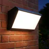 A modern black outdoor wall light with a bright white LED panel is mounted on a red brick wall at dusk. Produktbild-Vorschau
