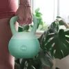 A person holding a green and white kettlebell in a home gym setting with plants in the background. Náhled obrázku produktu