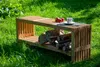 A wooden bench with a log storage compartment sits on a grassy lawn. Produktbild-Vorschau