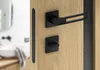 A modern black door handle set on a light wood door, with a blurred bathroom interior in the background. Produktbild-Vorschau