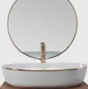A white oval basin with a gold rim and a gold faucet in front of a round mirror. Náhled obrázku produktu