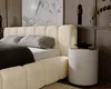 A close-up view of a modern beige upholstered bed with gray pillows, placed in a bedroom setting with wooden wall paneling and a marble-effect artwork. Náhled obrázku produktu