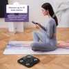 A woman is sitting on a yoga mat, looking at her phone, with a smart scale in the foreground. Produktbild-Vorschau