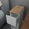 A close-up of a narrow white bedside table with an open drawer, next to a grey bed. The tabletop is made of wood. Náhled obrázku produktu