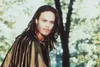 A man with long dark hair and a cape stands in a wooded area. Náhľad obrázku produktu