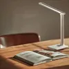 A desk lamp is positioned on a wooden table next to an open book. Produktbild-Vorschau