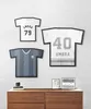 Three framed t-shirts are displayed on a white brick wall above a wooden dresser. Two framed pictures are on the dresser. Podgląd zdjęcia produktu