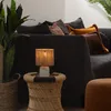 A close-up of a small table lamp with a woven shade sitting on a woven stool next to a sofa. Produktbild-Vorschau
