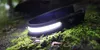 A black LED headlamp with a bright white light is resting on mossy ground in a forest setting. Náhled obrázku produktu