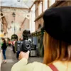 A person is holding a camera with a microphone attached, filming in an indoor market or arcade. Anteprima dell’immagine prodotto
