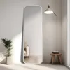 A full-length mirror stands against a white wall, reflecting a sofa and a floor lamp. A potted plant is to the left, and a small wooden stool with folded towels is to the right. Voir les images du produit