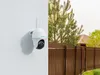 A security camera mounted on a white wall, with a brown fence and trees in the background. Produktbild-Vorschau