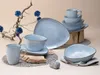 A collection of blue ceramic tableware, including plates, bowls, and mugs, arranged on a neutral background with decorative branches and stones. Náhľad obrázku produktu