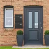 A front view of a modern brick house entrance with a dark gray door, a window, and two black mailboxes mounted on the wall. Two large planters with green bushes flank the doorway. Produktbild-Vorschau