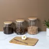 Three glass jars with wooden lids filled with coffee beans, dried fruit, and chickpeas, placed on a white marble countertop in front of a brown wall. A cutting board with a garlic bulb and a knife is in the foreground. Produktbild-Vorschau
