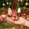 A hand holding a small Christmas gnome figurine, with other gnome figurines and Christmas tree branches in the background. Podgląd zdjęcia produktu