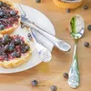 A close-up of a set of children's cutlery with a Jurassic World theme, placed on a wooden surface next to blueberry muffins and scattered blueberries. Produktbild-Vorschau