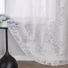 A close-up of a white sheer curtain with a gray floral pattern, draped near a window. Produktbild-Vorschau