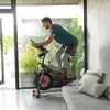 A man is exercising on an indoor cycling bike in a modern, well-lit room with large windows overlooking a garden. Produktbild-Vorschau