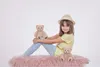 A young girl sits on a pink rug with two teddy bears. Voir les images du produit