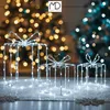 Three illuminated gift box decorations with white LED lights are arranged on a sparkly surface, with blurred Christmas trees in the background. Podgląd zdjęcia produktu